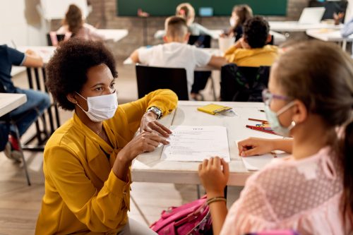 African American teacher and schoolgirl wearing protective face masks while discussing about test results on a class at the school.