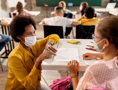 African American teacher and schoolgirl wearing protective face masks while discussing about test results on a class at the school.