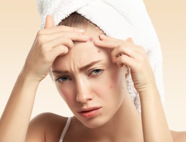 Headshot of displeased young blond woman with towel on her head, looking with painful face at the camera while squeezing pimple on her forehead. Portrait of Caucasian girl against blue wall background