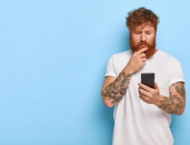 Serious bearded man looks attentively into screen, reads news online, updates software, has tattooes on arms, dressed in white casual t shirt, searches media files, stands indoor against blue wall