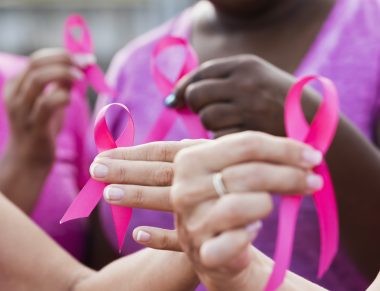 Cropped view of four multi-ethnic women wearing pink shirts, holding breast cancer awareness ribbons. The focus is on one of the hands.